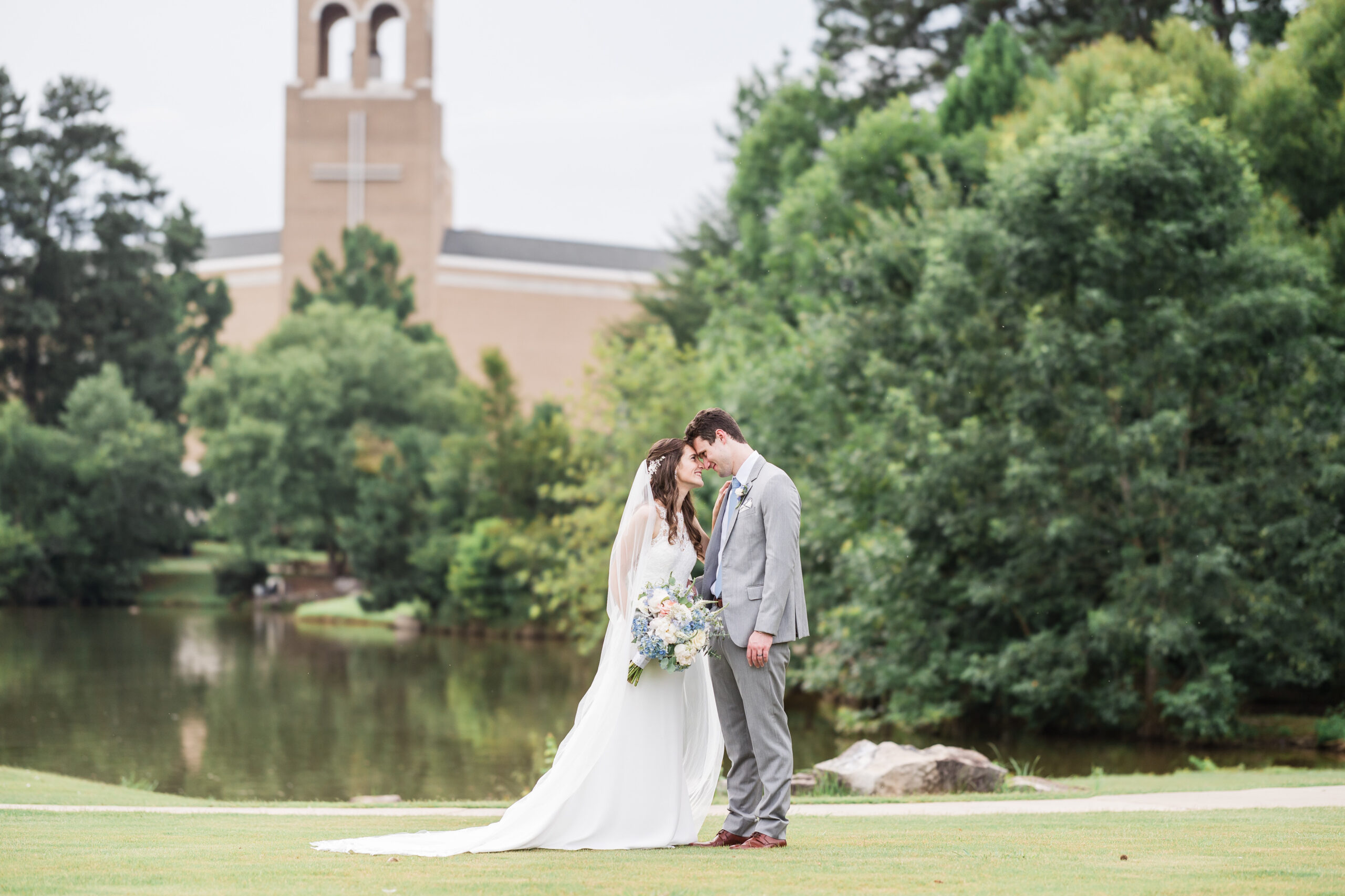 Bride and Groom put their heads together smiling with the church, lake, and the trees as their background