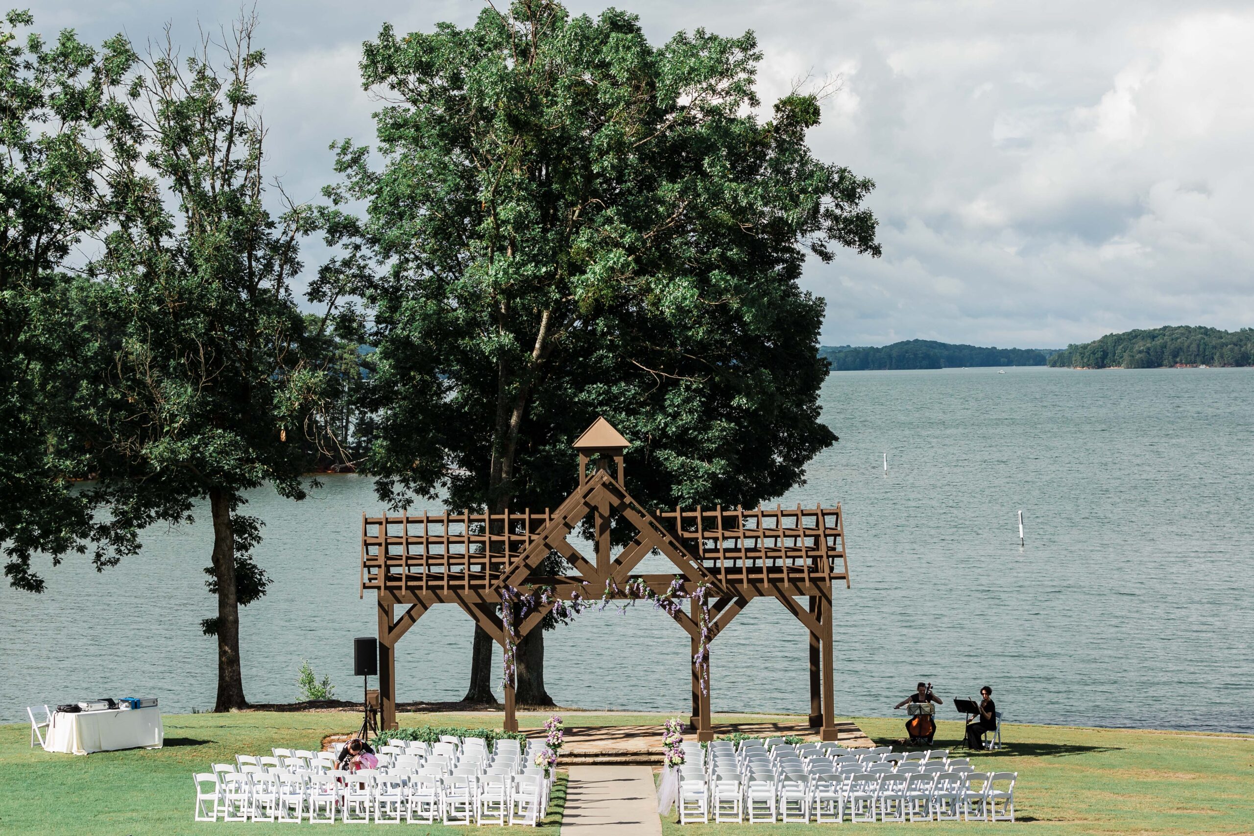 A brown gazebo and white chairs arranged neatly on a lakeshore with two musicians holding their instruments at the right side of the gazebo
