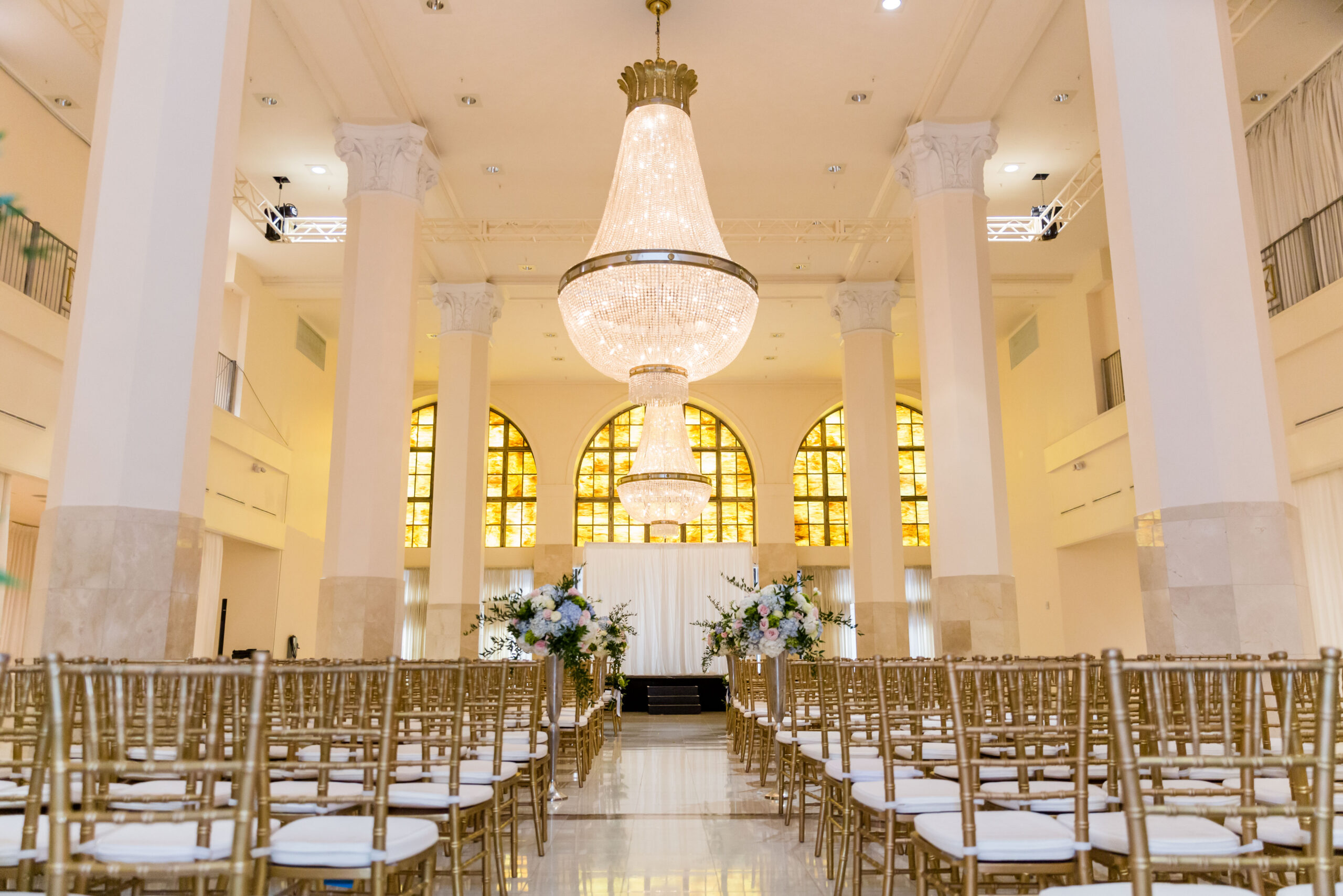 All white interiors inside a ballroom with four pillars and a grand chandelier in the middle and gold chairs piled neatly