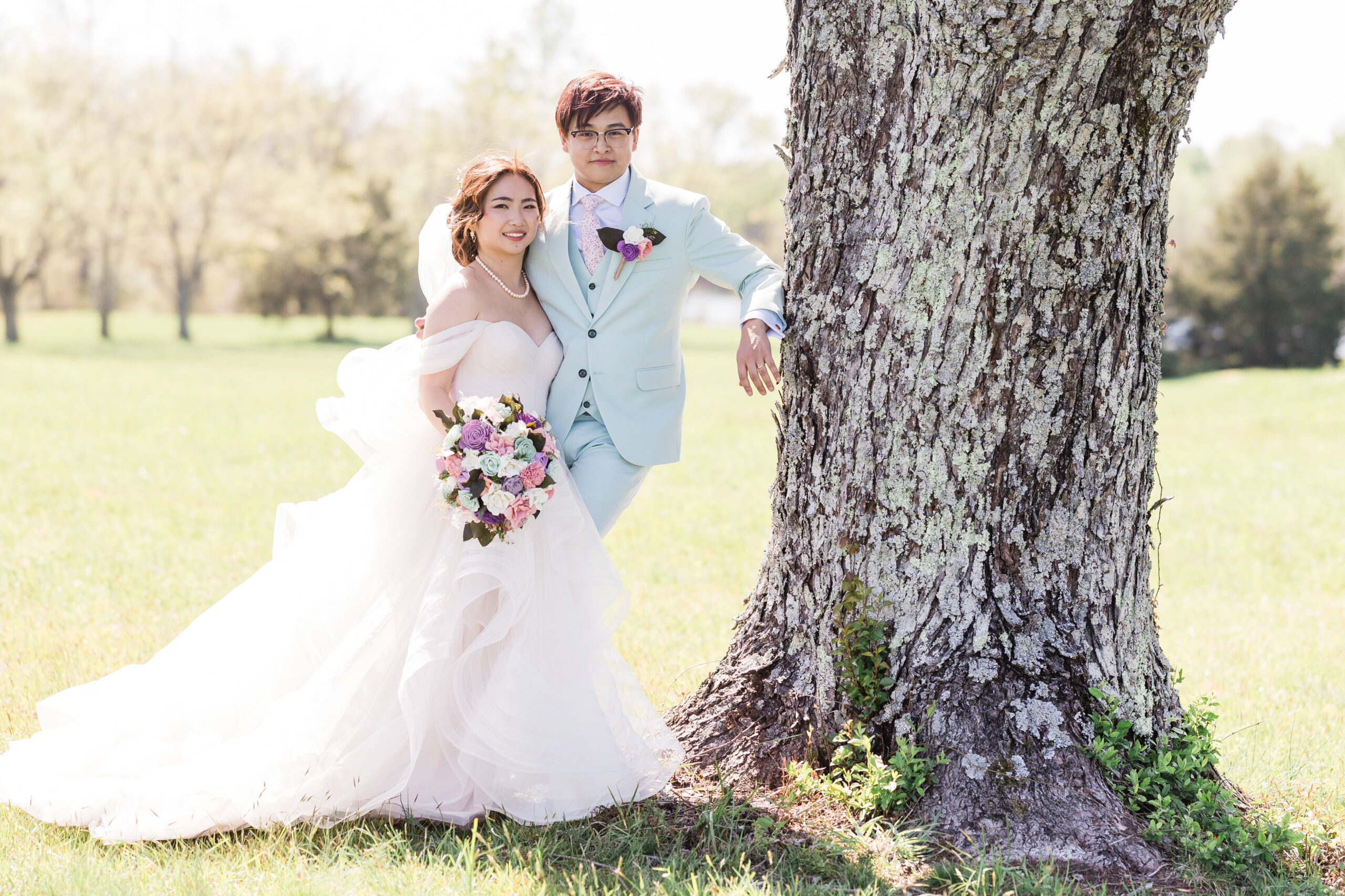 bride and groom stand beside each other as groom leans on a tree trunk and both of them smiling at the camera