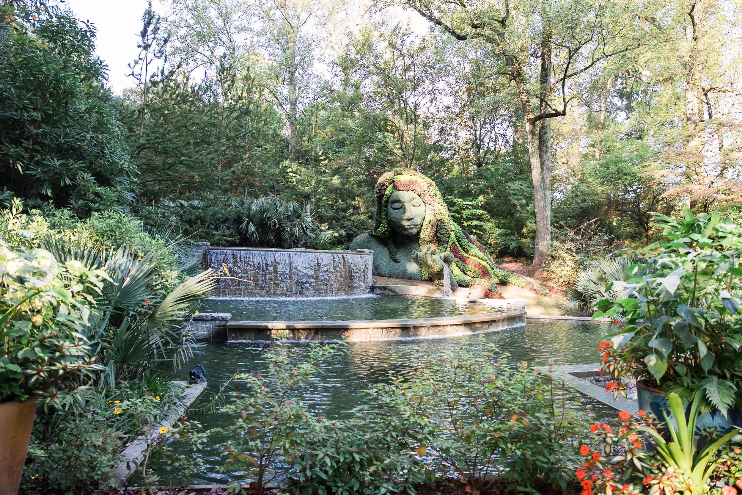 Photo of a garden with a waterfall that falls into a pond and a figure of a woman with leaves and flowers forming her face