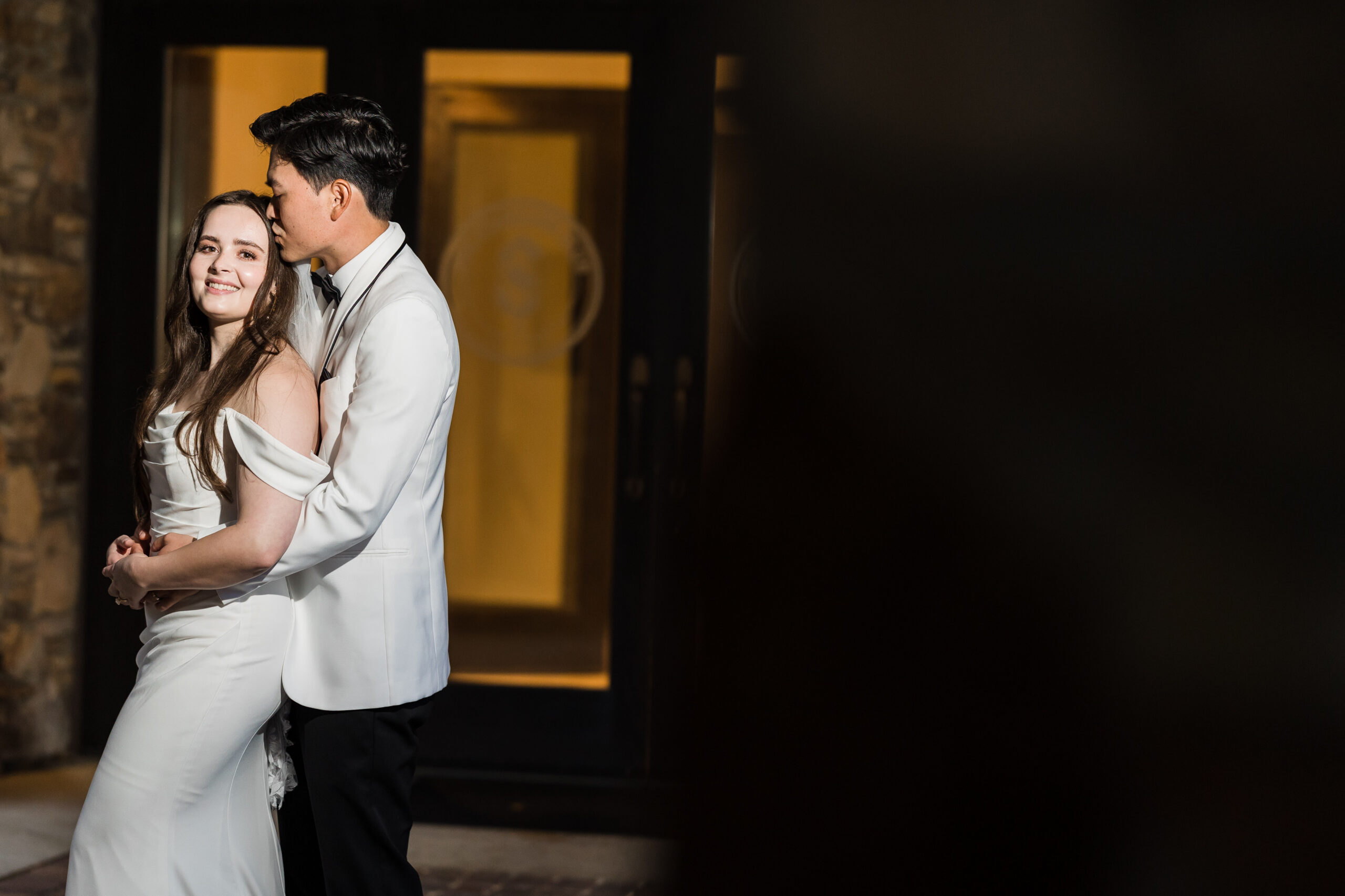 groom hugs bride from the back while kissing her forehead and she smiles at the camera